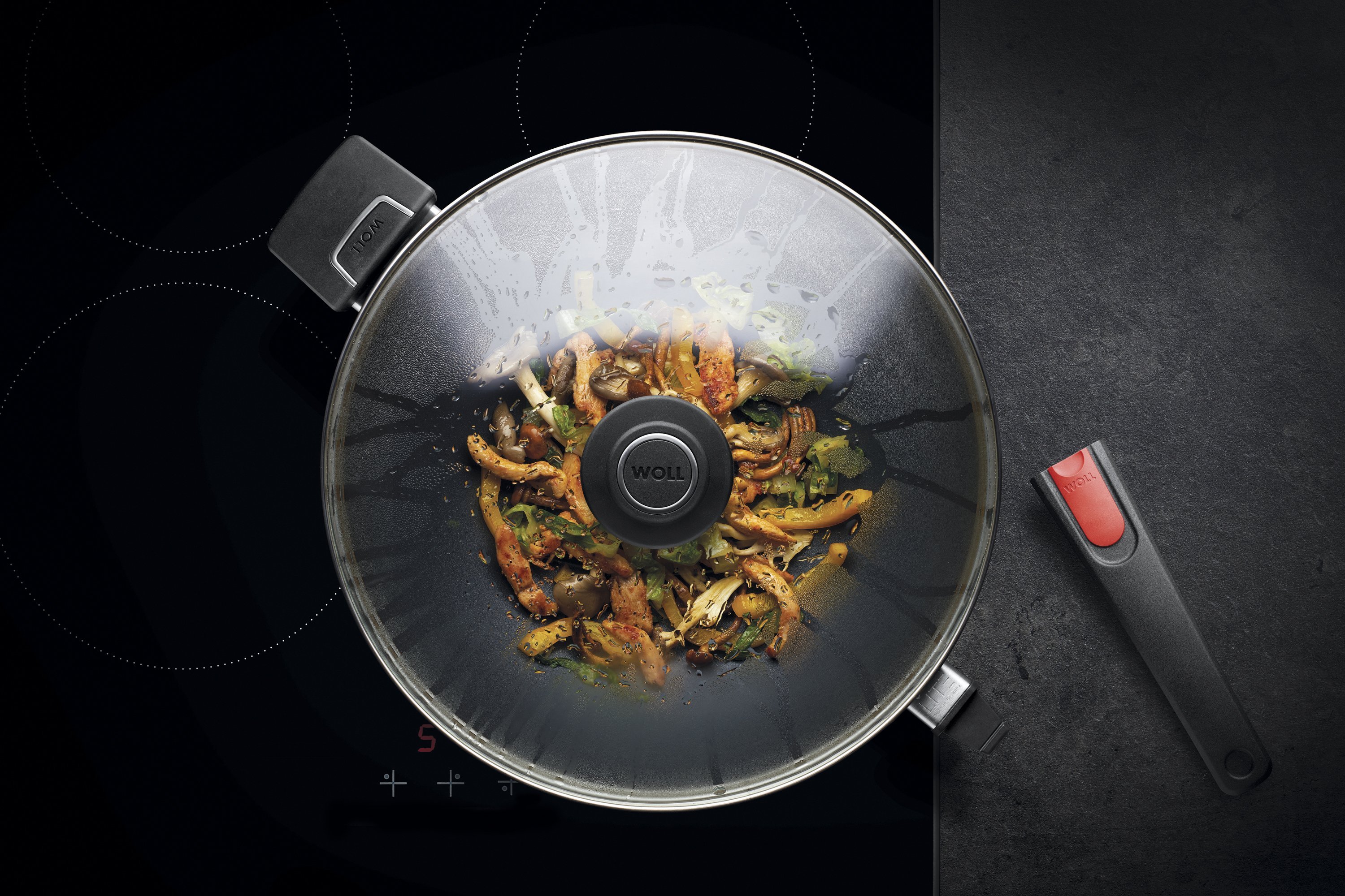 Auf dem Herd steht eine runde Diamond Lite Wok- und Rührpfanne In dieser befindet sich ein frisch zubereitetes Gericht. Am Glasdeckel hat sich Kondenswasser gebildet.