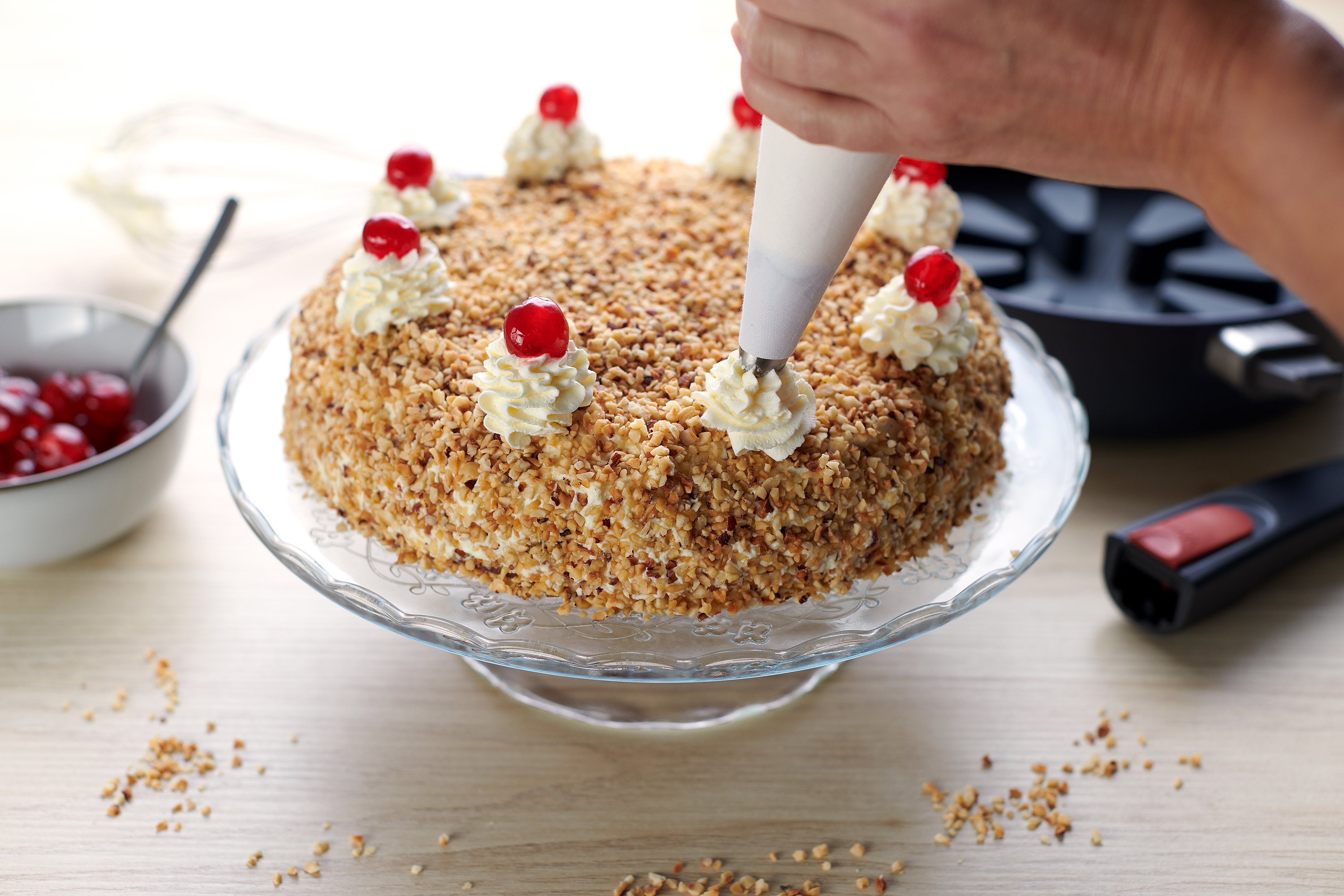 Ein Frankfurter Kranz wird mit Sahnehauben und glasierten Kirschen verziert. Im Hintergrund steht eine runde Diamond Lite Pfanne mit Silikon-Backeinsatz.
