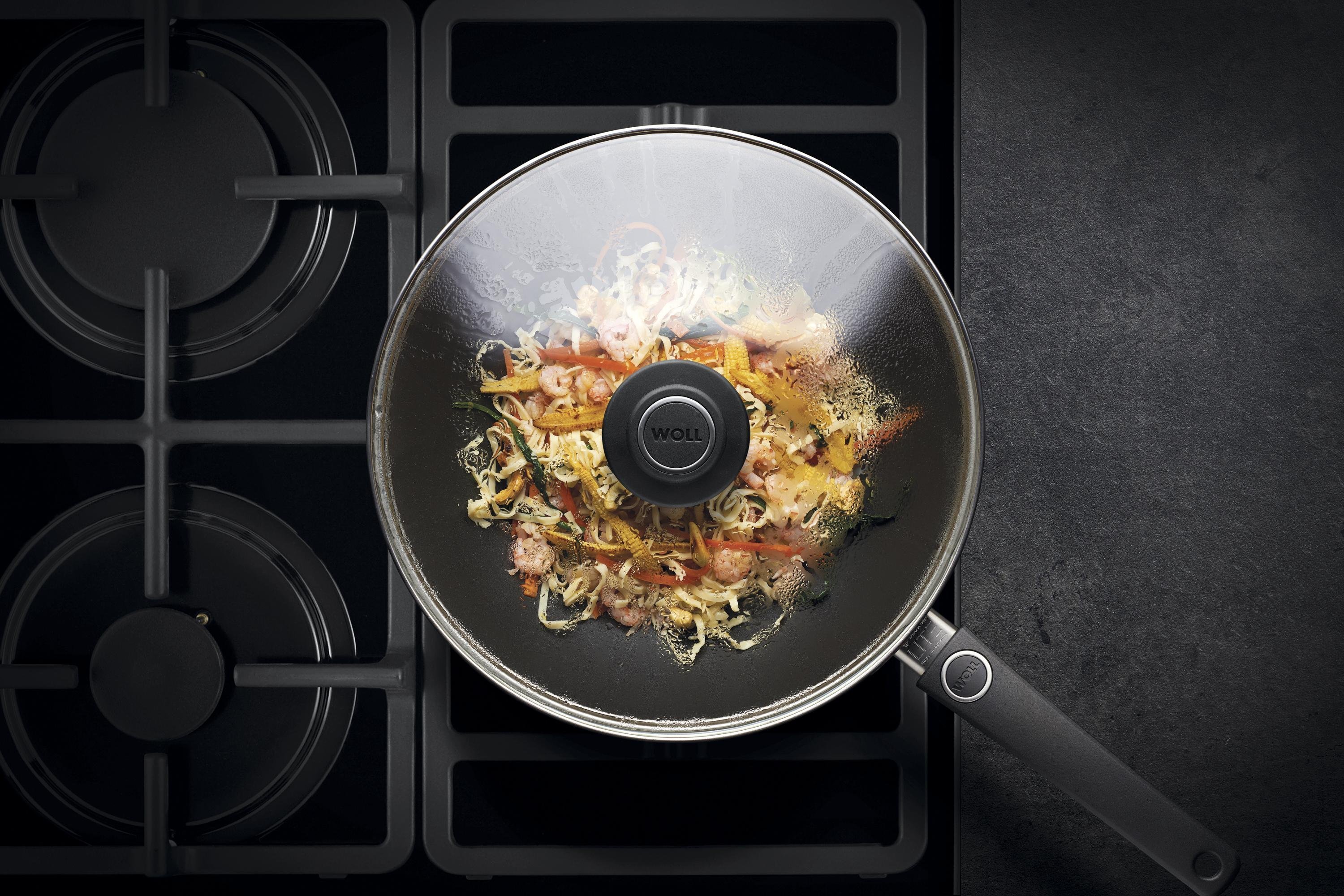 Auf dem Herd steht eine runde Diamond Lite Wok- und Rührpfanne In dieser befindet sich ein frisch zubereitetes Gericht. Am Glasdeckel hat sich Kondenswasser gebildet.