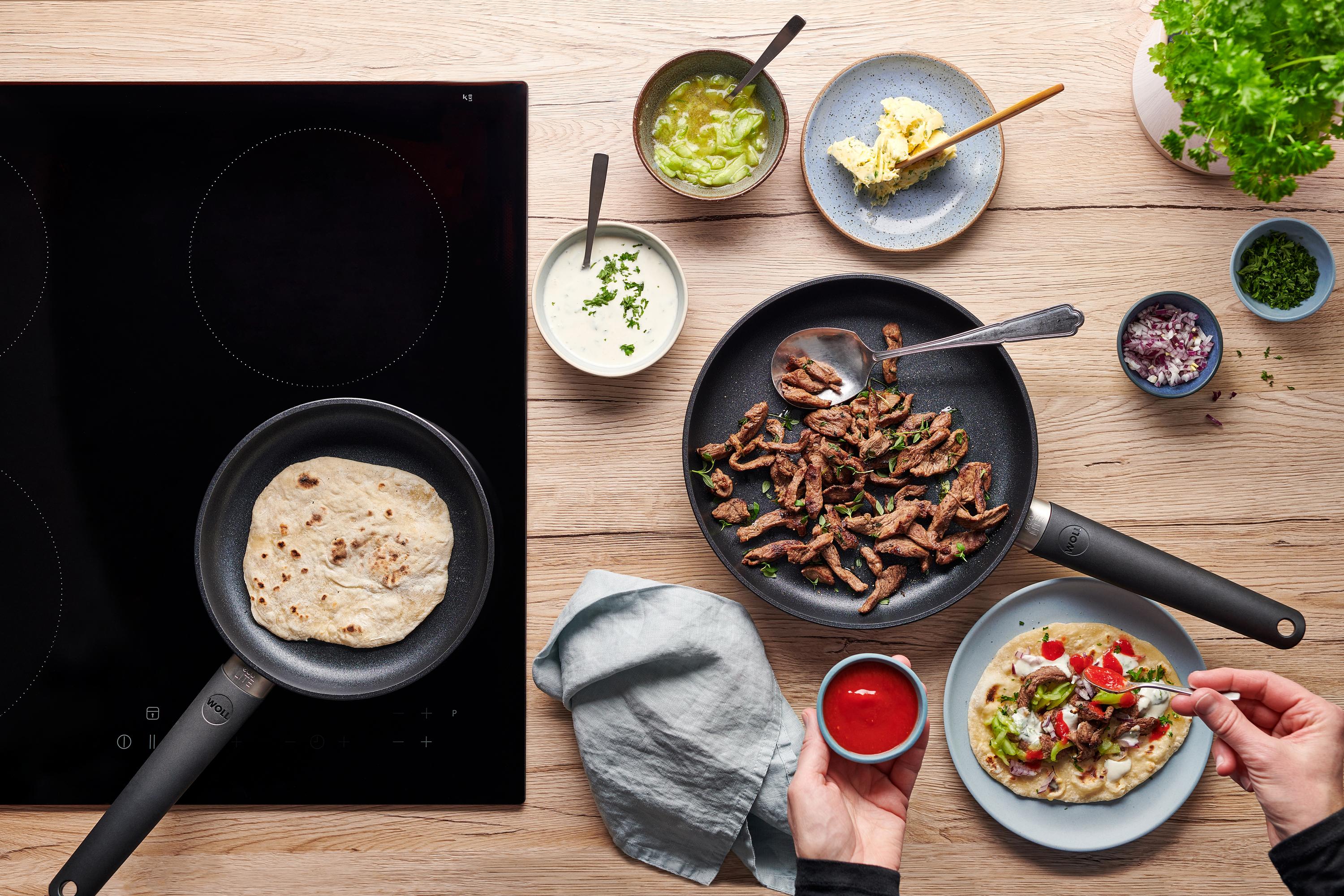 In der runden Eco-Lite-QXR-Pfanne werden Fladenbrote gebacken. Daneben liegt in einer Pfanne angebratenes Lamm und auf einem Teller ist Döner zubereitet.