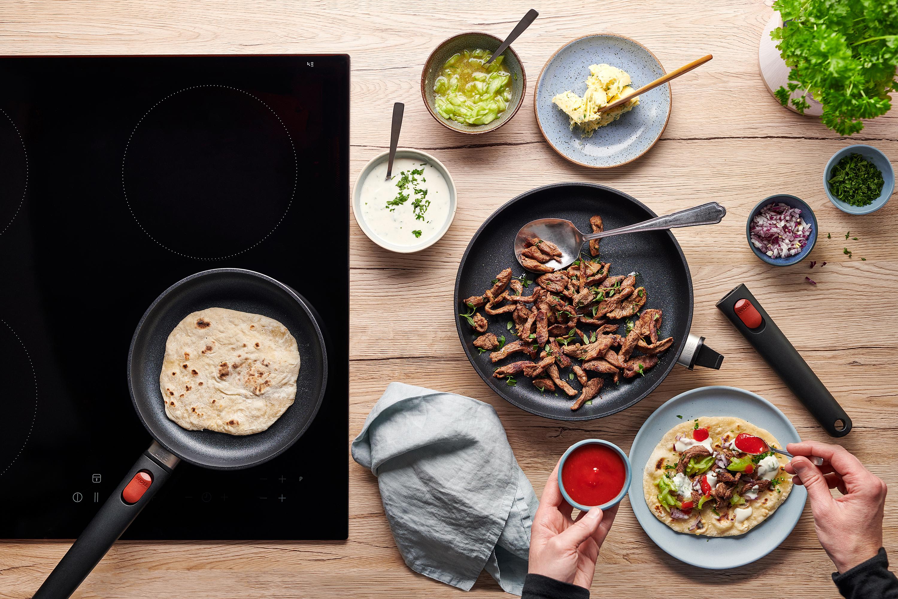 In der runden Eco-Lite-QXR-Pfanne werden Fladenbrote gebacken. Daneben liegt in einer Pfanne angebratenes Lamm und auf einem Teller ist Döner zubereitet.